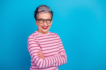Smiling woman wearing tiara and glasses in pink striped top standing confidently against bright blue background © deagreez