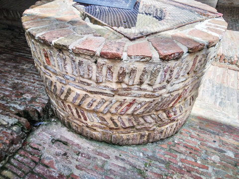 Ancient circular stone water well built with rustic terracotta bricks featuring a protective metal mesh grate situated in a historic cobblestone courtyard during the day