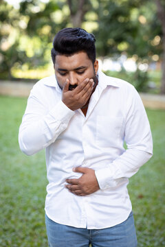 young adult south asian man in his 20s feeling nauseous and about to vomit in outdoors park, holding his stomach in distress