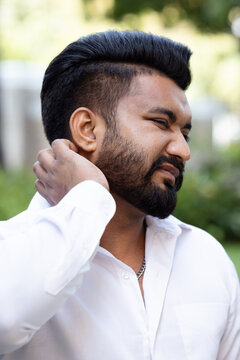 South asian 20s young adult man scratching his itchy neck from a mosquito bite or skin rash in outdoor green park scene, allergic reaction concept