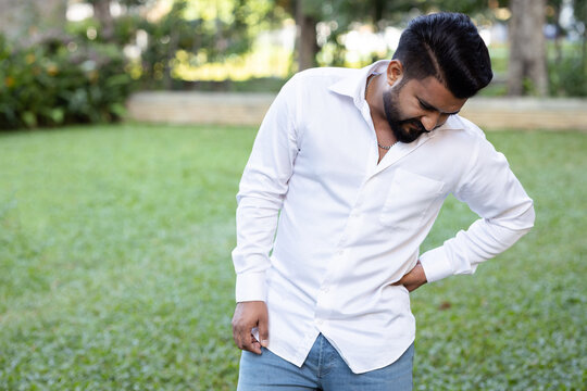 South Asian 20s young adult man suffering from waist and lower back pain, herniated disc, office syndrome, green park outdoor scene
