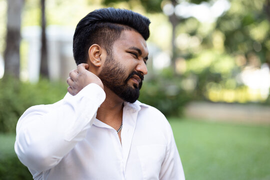 South asian 20s young adult man scratching his itchy neck from a mosquito bite or skin rash in outdoor green park scene, allergic reaction concept