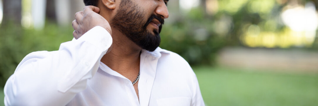 Hand closeup of South asian 20s young adult man scratching his itchy neck from a mosquito bite or skin rash in outdoor green park scene, allergic reaction concept