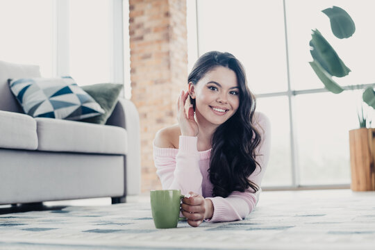 Smiling young woman relaxing on living room floor with coffee mug in cozy modern apartment enjoying a bright, leisurely morning
