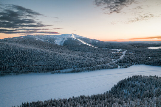 Aerial view of the snow-covered Levi ski resort and surrounding frozen landscape with dense pine forests and ski slopes under a soft sunset sky in Sirkka, Lapland, Finland.