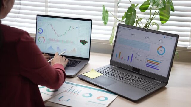 Businesswoman analyzing financial graphs and data on dual laptops at office desk