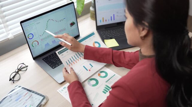 Businesswoman analyzing financial reports with charts on laptops and paper at her office desk
