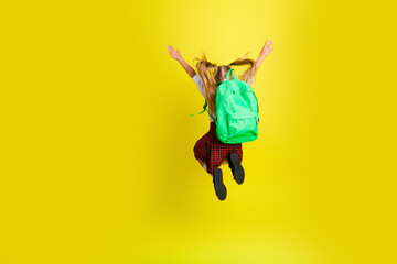 Obraz premium Schoolgirl jumping with green backpack against bright yellow background, joyful back-to-school moment celebrating childhood energy and excitement