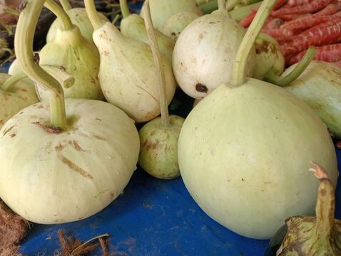 A close-up view of freshly harvested light green gourds displayed on a blue market stall surface, with other vegetables in the background.