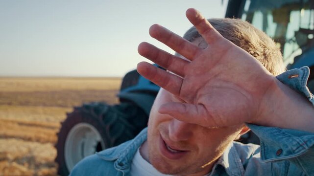 Young worker wipes sweat from forehead wiyh hand near tractor