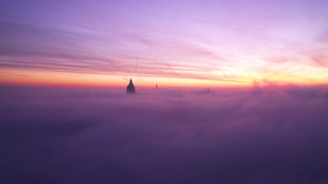 Aerial view of the Frankfurt skyline with a skyscraper piercing through a thick layer of fog during a vibrant sunset in Frankfurt am Main, Hessen, Germany.