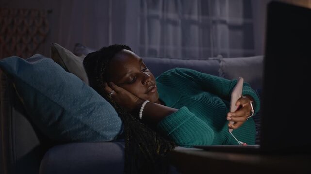 Young adult African American woman lying on sofa at night, checking smartphone and rubbing eyes in exhaustion while working on laptop. Footage represents digital burnout and remote work fatigue