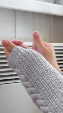 Close up of female palms touching electric convector heater. Vertical video. Woman in knitted sweater warming hands in cold room. Energy crisis, high utility bills. Cold home, winter poverty.