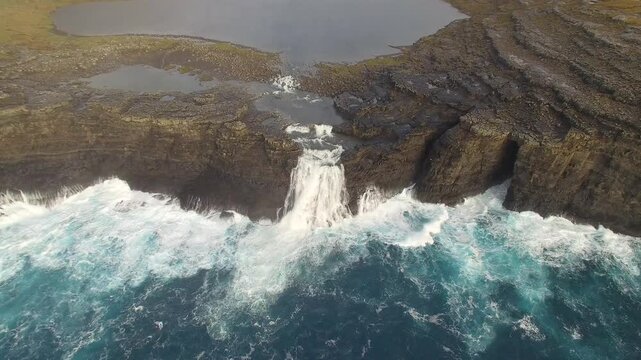 Aerial view of Mulafossur Waterfall cascading from a steep cliff into the turbulent North Atlantic Ocean with crashing waves and green grassy terrain in the Faroe Islands.