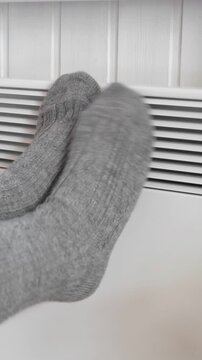 Person feet wearing thick wool socks leaning on radiator grid of electric convector heater. Vertical video. Warming in cold. Energy crisis, high utility bills. Winter house freezing. High heating cost
