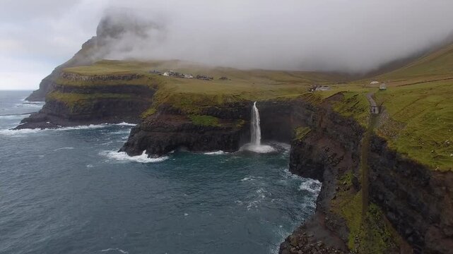 Aerial view of Mulafossur waterfall cascading down steep cliffs into the Atlantic Ocean, surrounded by green grassy slopes and misty clouds in the Faroe Islands.