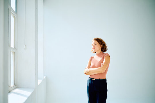 Woman standing with crossed arms, looking out a bright window, deeply thinking and developing new business ideas