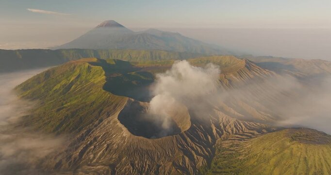 Aerial view of Mount Bromo and Tengger Caldera with volcanic smoke rising from the crater, surrounded by lush green slopes and misty caldera landscape, Indonesia.