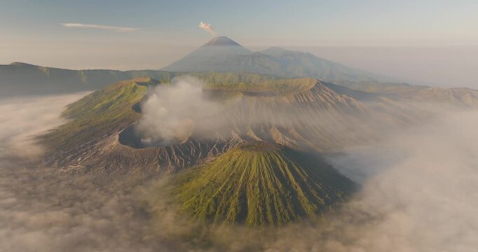 Aerial view of Mount Bromo and Tengger Caldera with volcanic smoke and mist over green slopes at sunrise in Indonesia.