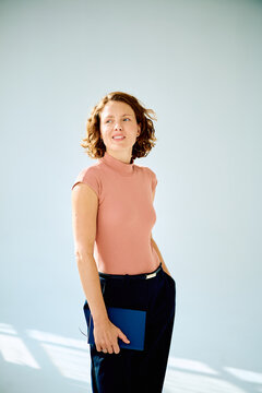 Professional young woman standing against light background, holding a notebook, and looking to the side with a gentle smile