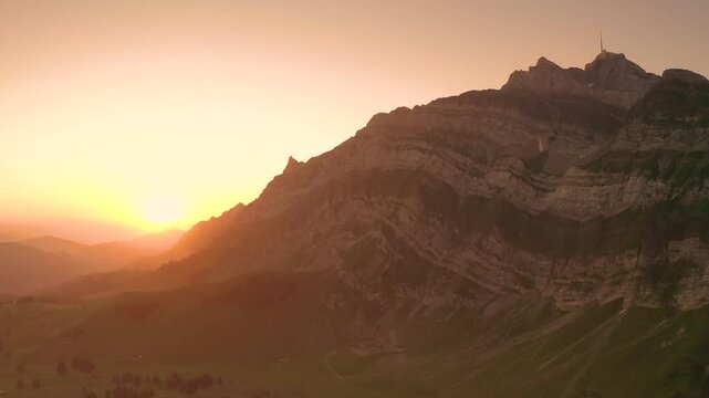 Aerial view of the rocky Alpstein mountain range illuminated by the golden sunset light over the green slopes in Appenzell, Appenzell Innerrhoden, Switzerland.