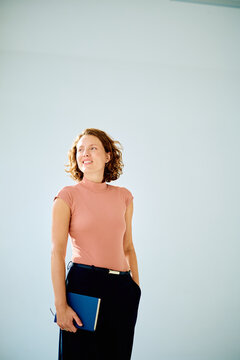 Businesswoman standing in a studio, wearing a casual shirt and holding a blue notebook, smiling and looking to the side