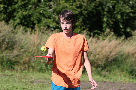 Young player balancing a tennis ball on a red racket, practicing keepie uppie skills in the countryside