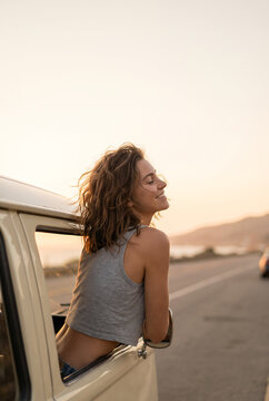 Young woman enjoying sunset breeze from car window on desert highway