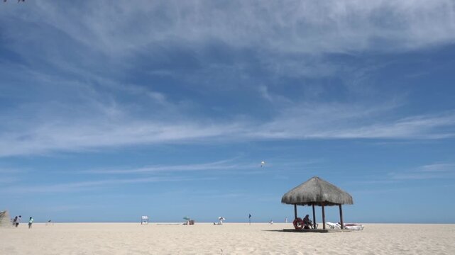 A serene tropical beach scene with a thatched roof hut on white sand under a vast blue sky with wispy clouds and distant parasailer.