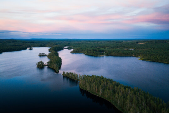 Aerial view of Hossa National Park featuring forested islands and peninsulas stretching across a calm lake under a soft pink and blue twilight sky in Ruhtinansalmi, Kainuu, Finland.