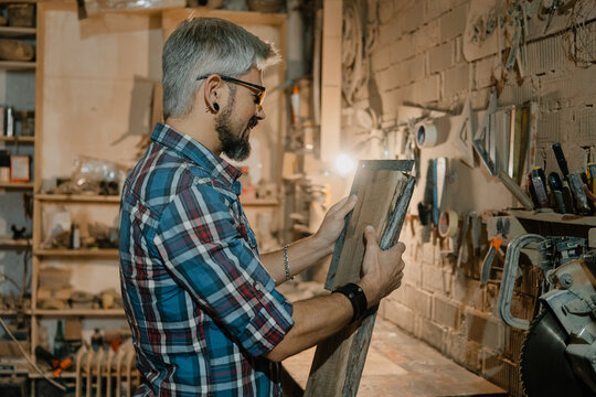 Carpenter examining reclaimed wood plank in a busy woodworking workshop with tools and sawdust