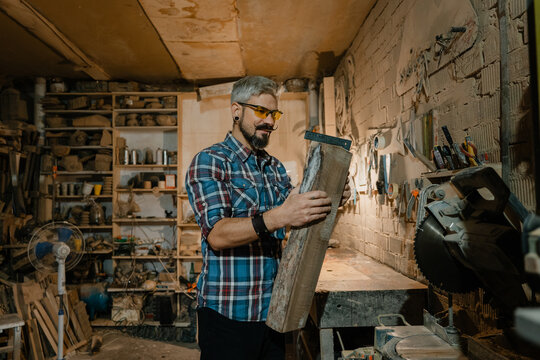 Carpenter inspecting a piece of reclaimed wood in a cluttered woodworking workshop wearing safety glasses