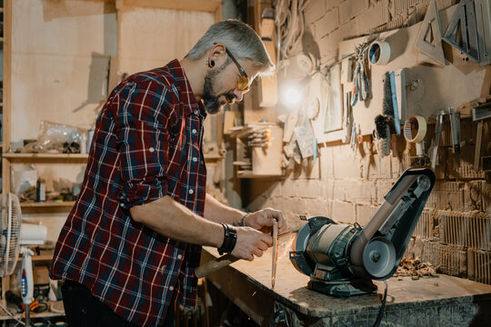 Middle-aged craftsman sharpening a chisel at a bench grinder in a cluttered woodworking workshop