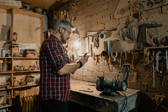 Bearded craftsman working with tools in a rustic woodworking workshop lit by a single lamp