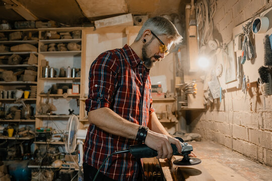 Carpenter sanding a wooden workpiece with a power sander in a cluttered workshop