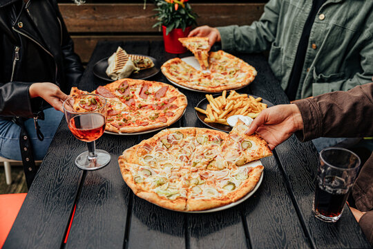 Group of diners share assorted pizzas, French fries, grilled wrap and drinks around a dark outdoor restaurant table, creating a casual social dining scene