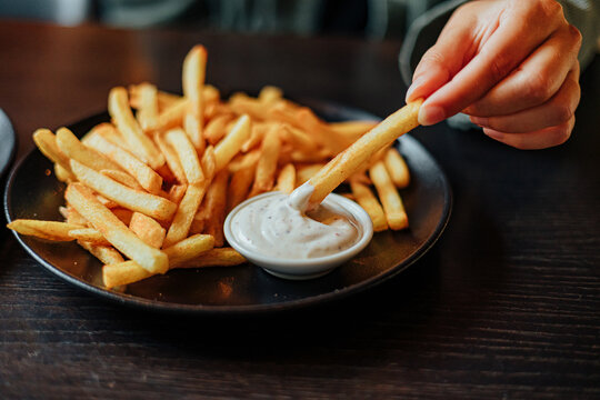 Close-up of a hand dipping a golden French fry into creamy seasoned sauce beside a plate piled with crispy fries at a restaurant table