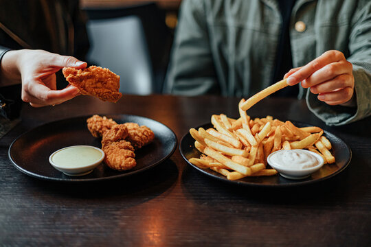 Close-up of two diners holding crispy fried chicken and a French fry beside plates of fries, dipping sauces and chicken pieces at a restaurant table