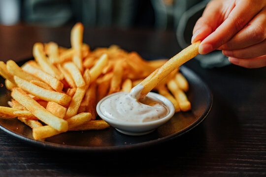Close-up of a hand dipping a golden French fry into creamy seasoned sauce beside a plate piled with crispy fries at a restaurant table