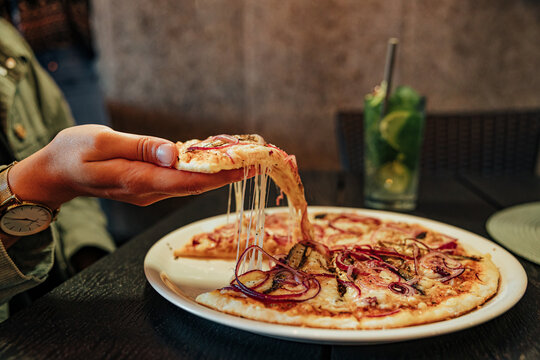 Close-up of a diner lifting a hot pizza slice with stretchy melted cheese and red onion toppings, with a mint drink in the background