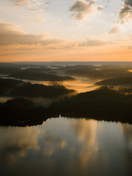 Aerial view of a serene lake surrounded by dense pine forests and rolling hills covered in morning mist under a soft golden sky in Helsinki, Uusimaa, Finland.