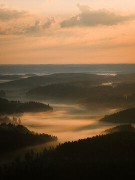 Aerial view of the misty landscape of Hiidenvesi at sunrise with dense pine forests and rolling hills under a golden sky in Helsinki, Uusimaa, Finland.