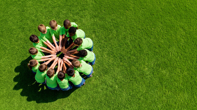Diverse group of children sitting in a circle on grass with hands stacked together showing teamwork and unity
