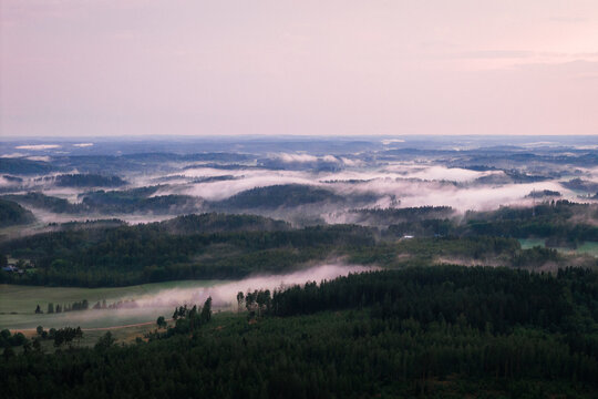 Aerial view of the misty landscape around Hiidenvesi lake, featuring dense evergreen forests, rolling hills, and low-hanging fog in Helsinki, Uusimaa, Finland.