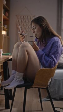 Vertical video of young adult Caucasian woman eating meal from bowl while staring at smartphone screen. Footage illustrating digital addiction and screen time habits for mental health campaigns