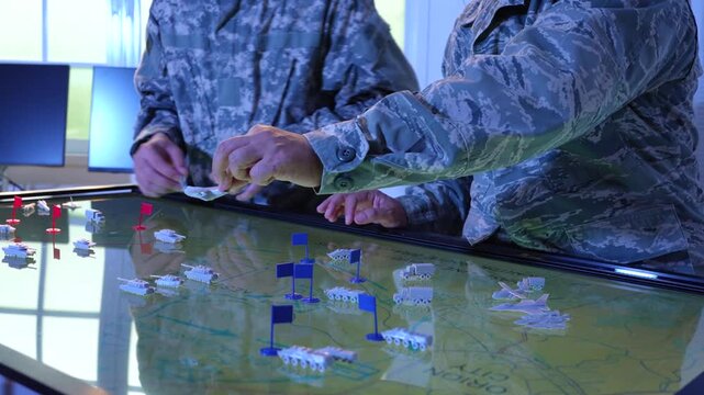 Military personnel in camouflage uniforms use an interactive digital map table to plan a tactical operation with unit markers.