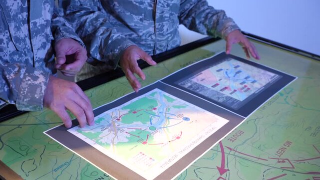 Military personnel in camouflage uniforms study printed maps on a glowing digital table during a tactical planning briefing.