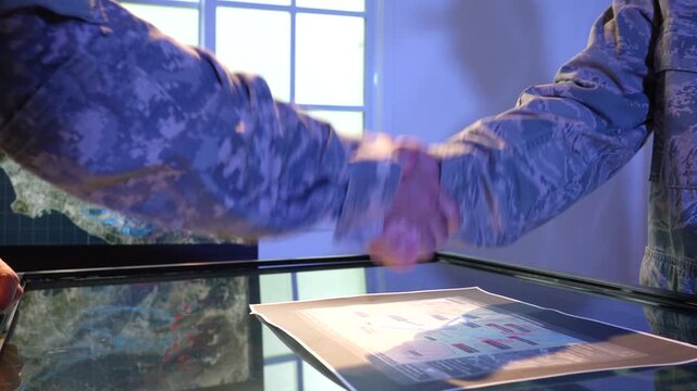 Two military personnel shake hands over a glass table with a map and documents, suggesting cooperation, planning, and strategic agreement.