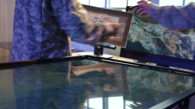 Analyst points at a digital map on a large touch screen beside computer monitors, geographic data in a modern control room.