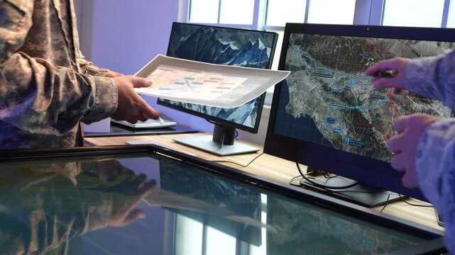 Military personnel use digital maps on a touchscreen table and computer monitors during a command center planning session.